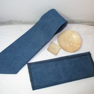 May include: A denim blue necktie, pocket square, and two buttons. The tie is folded and lies next to the pocket square. A large, round, wooden button with four holes and a square button with two holes are also present.