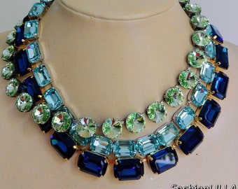 May include: A multi-layered statement necklace featuring large, faceted, rectangular crystals in shades of blue and green. The necklace is made of gold-toned metal and has a graduated design with the largest crystals at the bottom.
