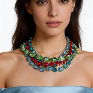 Könnte beinhalten: Eine mehrreihige Halskette mit einer lebendigen Auswahl an farbigen Edelsteinen. Die Halskette zeigt eine Mischung aus grünen, roten und blauen Steinen in verschiedenen Formen, was ein auffälliges Accessoire ergibt. Die Edelsteine sind in einem silberfarbenen Metall gefasst.