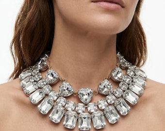 May include: A multi-layered statement necklace featuring numerous clear, faceted rectangular and triangular crystals set in a gold-toned metal frame. The design consists of three distinct rows of stones that drape around the neck, creating a dense, reflective, and high-shine appearance with a secure clasp connection.