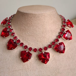 Può includere: Una collana abbagliante con grandi pietre preziose rosse a forma di cuore. La collana ha una catena argentata con pietre rosse rotonde più piccole. Le pietre preziose sono sfaccettate, catturando la luce e creando un effetto scintillante.