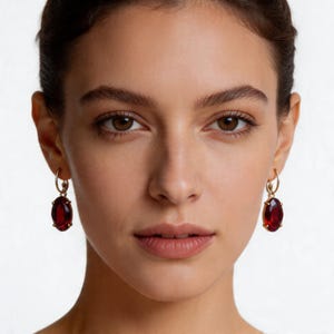 May include: A close-up shot of a person wearing gold hoop earrings with large, oval, deep red gemstones. The earrings are the focal point, set against the person's skin and hair.