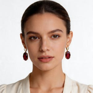 May include: Gold-tone hoop earrings with large, oval, deep red gemstones. The earrings are a classic design, featuring a polished gold finish and a vibrant, eye-catching color.