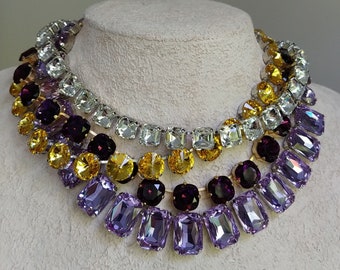 May include: A multi-layered necklace with a variety of crystal colors. The necklace features a bottom layer of lavender rectangular crystals, a middle layer of yellow and gold round crystals, and a top layer of clear, white, and silver square crystals.