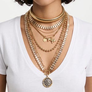 Peut inclure: Un ensemble de colliers dorés superposés comprenant une chaîne serpent épaisse, une chaîne à maillons rectangulaires, une chaîne fine avec une breloque épingle à nourrice, une chaîne perlée et une longue chaîne avec des cristaux baguette.
