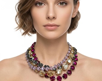 May include: A multi-strand statement necklace featuring faceted crystals in shades of magenta, deep purple, amber, and charcoal gray. The necklace consists of several layered rows of round and rectangular cut stones set in a metallic frame.