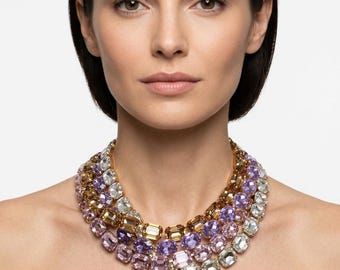 May include: A multi-strand statement necklace featuring rows of faceted rectangular crystals in shades of clear, light pink, lavender, and amber. The stones are set in a gold-tone metal frame, creating a layered, collar-style design that rests against the neck.
