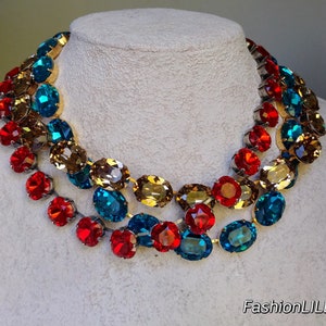 May include: A multi-strand necklace featuring large oval-shaped faceted gemstones in shades of red, amber, and teal. The necklace is crafted from silver-toned metal.