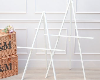 Può includere: Due cavalletti in legno bianco con barre orizzontali per esporre opere d'arte o cartelli. Sono posizionati contro un muro bianco con modanature decorative. A sinistra c'è un cestino di vimini con fiori.