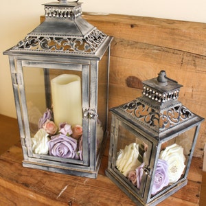 May include: Two decorative lanterns with glass panels and ornate metal frames. The larger lantern contains a cream-coloured candle and floral arrangement of lavender, pink, and white roses. The smaller lantern has a similar arrangement. Both sit on a wooden surface.