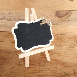 May include: A small wooden easel with a black chalkboard sign. The chalkboard has a white border and a decorative edge. The easel is made of natural wood and has a rustic look.