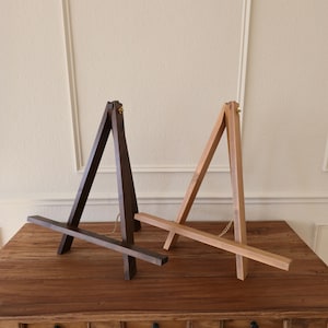 Puede incluir: Dos caballetes de madera, uno marrón oscuro y otro marrón claro, se exhiben sobre una superficie de madera. Cada caballete tiene un marco triangular con una barra de soporte horizontal. Una pequeña cadena conecta la parte superior de cada caballete.