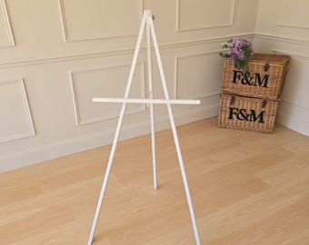 Puede incluir: Un caballete de madera blanco con un diseño de tres patas. El caballete tiene una barra horizontal para sostener un lienzo o una obra de arte. El fondo incluye dos cestas de mimbre con el texto "F&M" y un ramo de flores moradas.