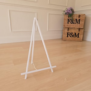 Può includere: Un cavalletto di legno bianco con un telaio triangolare è appoggiato su un pavimento in legno chiaro. Il cavalletto ha un design semplice con una catena per fissare le gambe. Sullo sfondo ci sono due cesti di vimini con le lettere "F&M" e un mazzo di fiori viola.