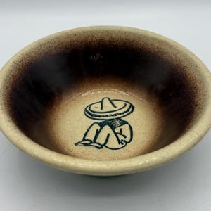 May include: A round ceramic bowl with a speckled cream-colored exterior and interior. The interior features a dark brown gradient rim and a dark blue line drawing of a person wearing a wide-brimmed hat sitting on the bottom of the bowl.