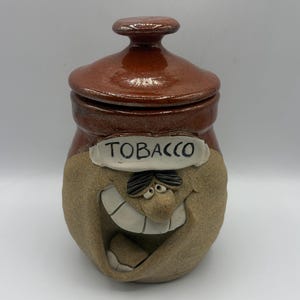 May include: A ceramic tobacco jar with a glossy brown lid and a textured tan base. The jar features a sculpted, smiling face with prominent white teeth and dark hair. A white label with the black text TOBACCO is centered above the face.