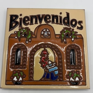 May include: A ceramic tile with the word "Bienvenidos" in brown lettering. The tile features a brown building design with a figure and decorative elements in green, red, and white. The tile is rectangular.