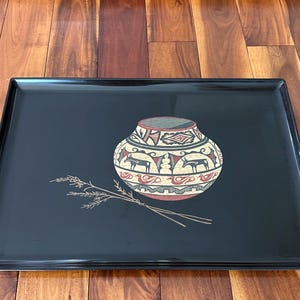 May include: A rectangular black serving tray with a decorative design. The tray features a central image of a pottery vessel with red, tan, and white patterns, along with a sprig of wheat. The tray is likely made of a durable material.