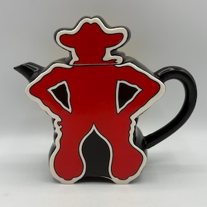 May include: A black ceramic teapot with a red figure design. The figure is outlined in white and has a cowboy hat. The handle and spout are black. The teapot is on a white background.