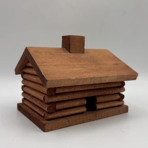 May include: A small, brown wooden model of a log cabin featuring a gabled roof, a central chimney, and horizontal log-style walls. The structure is built on a flat rectangular base with a small opening representing a doorway on the front side.