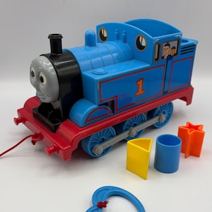 May include: A blue plastic toy train engine with a grey face, black smokestack, and the number 1 on the side. It features a red base, a red pull string with a blue handle, and three plastic shape-sorting blocks in yellow, blue, and orange.