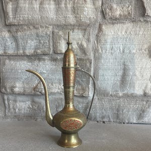 May include: An antique brass pitcher with a long, curved spout and a decorative handle. The pitcher features intricate red and gold etched designs. The base is round and the top has a pointed finial. The pitcher is likely used for serving beverages.