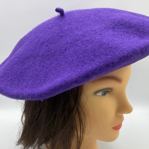 May include: A vibrant purple beret made of felt. The hat is round with a small, rounded stem on top. The beret is displayed on a mannequin head with brown hair and a neutral skin tone. The beret is a classic fashion accessory.