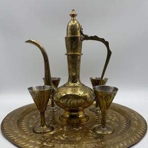 May include: A vintage brass tea set, including a decorative pitcher, small goblets, and a round tray. The set features intricate floral patterns and a warm, golden hue. The pitcher has a curved spout and handle.