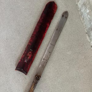 May include: A vintage knife with a long, silver blade and a textured, brown handle. The knife is next to a red leather sheath. The knife and sheath are on a gray surface.