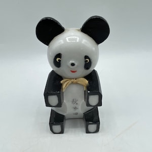 May include: A black and white ceramic panda figurine with a gold bow. The panda is sitting with its paws in front of it. The figurine has a black base and black ears. The panda has a black nose and black eyes with yellow pupils. The panda has a small red mouth. The figurine has Japanese writing on the bottom.