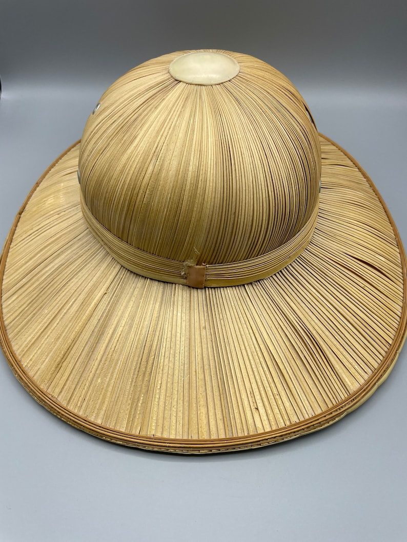 Puede incluir: Un sombrero de paja de ala ancha con una corona redonda. El sombrero est&aacute; hecho de paja tejida y tiene un color marr&oacute;n claro. La corona tiene un detalle circular blanco en la parte superior.