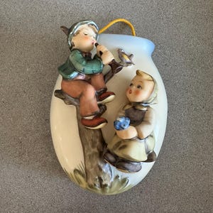 May include: A ceramic wall hanging featuring two children. One child sits on a tree branch playing a flute, while the other holds flowers. The background is a vase shape with a yellow string for hanging. The children are wearing green, brown, and yellow clothing.