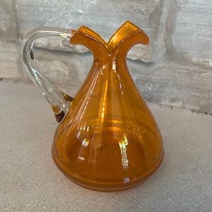 May include: An amber-colored glass pitcher with a clear glass handle. The pitcher has a unique, flared top and a rounded base. The handle curves gracefully from the top to the side of the pitcher. The pitcher is sitting on a gray surface.
