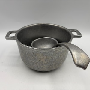May include: A gray metal cooking pot with two handles and a matching ladle. The pot has a rounded shape and a matte finish. The ladle rests inside the pot, with its handle extending over the rim.