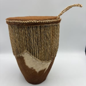 May include: A handcrafted drum with a brown, textured hide top and a woven handle. The body features a fringe of natural fibers and a lower section of brown and white patterned hide. This unique piece is a musical instrument.