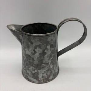 May include: A galvanized metal pitcher with a spout and a curved handle. The pitcher has a mottled gray finish and a cylindrical body. The handle is attached to the side of the pitcher. The pitcher is likely used for decorative purposes.
