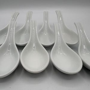 May include: A collection of seven white ceramic soup spoons. Each spoon has a deep, rounded bowl and a long, slender handle. The spoons are arranged in a slightly overlapping pattern, showcasing their clean, minimalist design.