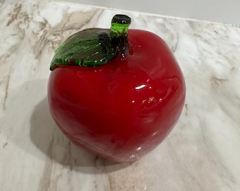 May include: A glossy, red glass apple with a green stem and leaf. The apple is a vibrant shade of red, with a smooth, reflective surface. The stem and leaf are a translucent green, adding contrast to the design. A decorative piece.