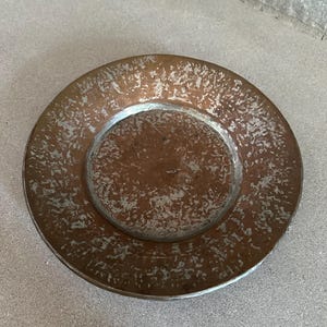 May include: A round, copper-colored plate with a textured surface and a silver-colored rim. The plate has a rustic, aged appearance, suggesting it may be vintage or handmade. The plate is suitable for serving food or as a decorative item.