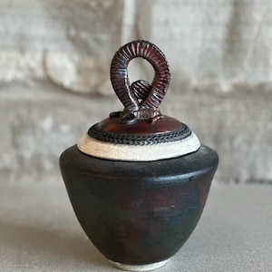 May include: A handcrafted ceramic jar with a dark, mottled finish. The lid features a loop handle and a decorative white band with a woven pattern. The jar's body is a deep, earthy tone, with a white base.