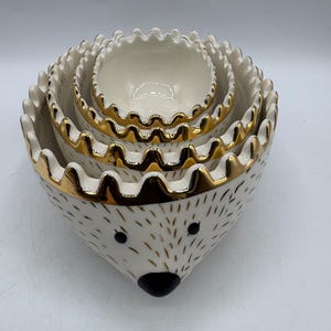 May include: A set of four nested ceramic bowls shaped like a hedgehog. The bowls are white with gold-painted scalloped rims, black painted eyes, a black nose, and small gold dash markings representing quills. Each bowl features text on the inner rim.