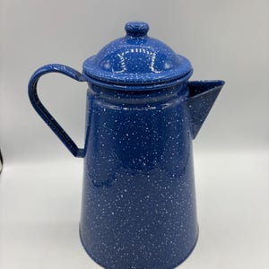 May include: A tall, cylindrical blue enamel coffee pot featuring a white speckled pattern throughout the exterior. The pot includes a matching lid with a small knob handle, a curved side handle, and a pointed pouring spout on the opposite side.