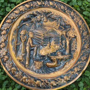 May include: A round, bronze-colored decorative plate with a detailed relief scene of a log cabin and figures. The plate's border features an acorn and leaf design. The central scene depicts a man, woman, and a cabin.