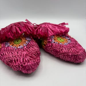 May include: A pair of bright pink woven slippers with a colorful floral design. The slippers have a fringe detail at the top and are made of a woven material. The floral design includes orange, green, and purple accents.
