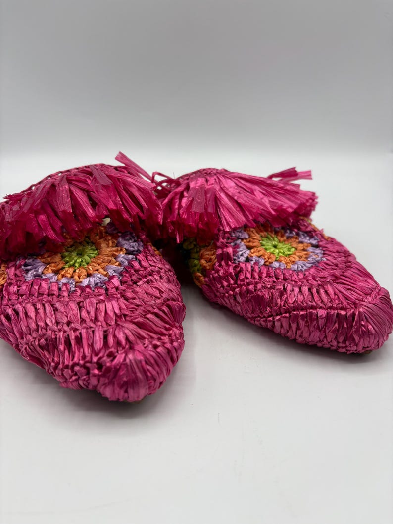 May include: A pair of bright pink woven slippers with a colorful floral design. The slippers have a fringe detail at the top and are made of a woven material. The floral design includes orange, green, and purple accents.