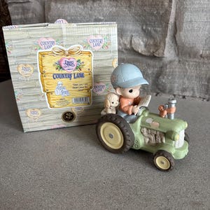 May include: A Precious Moments figurine of a boy driving a green tractor with a small dog passenger. The figurine is in front of a box with the words "Country Lane" and "Dear Jon I Will Never Leave You" on it. The figurine is on a gray surface.