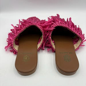 May include: A pair of bright pink raffia slip-on sandals. The sandals have a brown sole and a fringed upper. The interior of the shoe is a light beige color. The brand name "DE SENA" is printed on the insole.