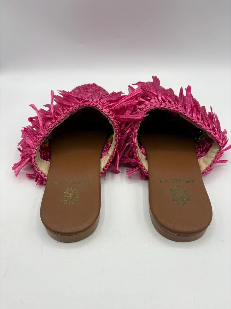 May include: A pair of bright pink raffia slip-on sandals. The sandals have a brown sole and a fringed upper. The interior of the shoe is a light beige color. The brand name "DE SENA" is printed on the insole.