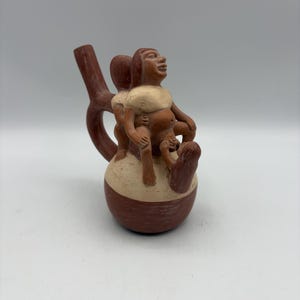 May include: A terracotta ceramic vessel featuring a figural design. The vessel has a bulbous base in brown and tan, with a spout and handle. Three figures are sculpted on top, with one looking upwards.