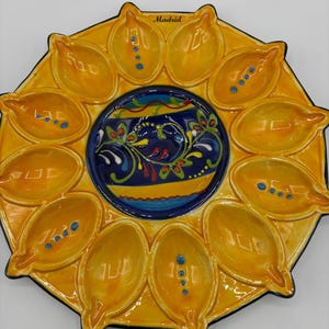 May include: A round ceramic serving platter with a central dark blue circular section featuring colorful floral patterns. Surrounding the center are twelve yellow, leaf-shaped compartments, each with small blue dots. The word Madrid is printed at the top.